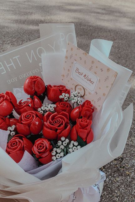 A bouquet of vibrant red roses and small white filler flowers arranged together, wrapped in white and semi-transparent paper. The bouquet is accompanied by a small card with the inscription 'Best Wish