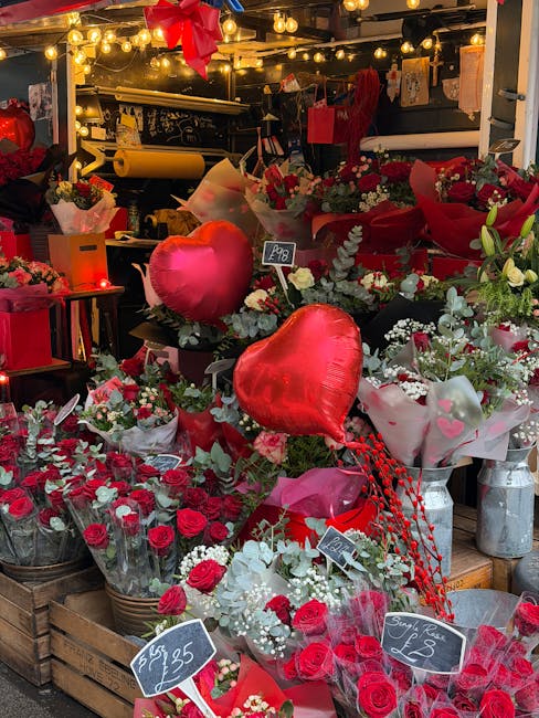 An indoor floral display featuring numerous bouquets and arrangements predominantly composed of red and white flowers, including roses and lilies. The arrangement is accentuated with decorative heart-