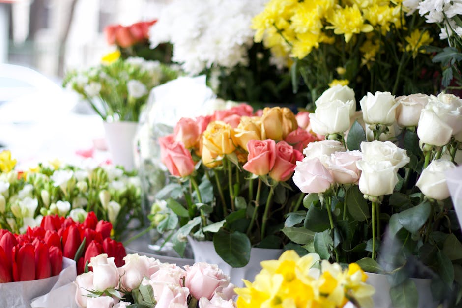 A vibrant display of fresh roses and tulips arranged in bouquets at an outdoor flower stall. The roses are in shades of white, pink, yellow, and peach, with lush green leaves and stems. The tulips are