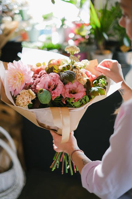 A person holding a large bouquet of fresh flowers, including soft pink peonies, white blossoms, and green foliage, wrapped in cream-colored paper with a satin ribbon. The bouquet is arranged in a lush