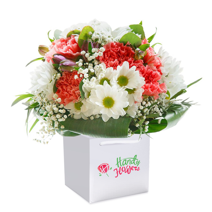 Presented upright in a flawless square white gift bag, this bouquet captures the lushness and vitality of early spring in Old Oak Common, much like a walk past the greenery of Wormwood Scrubs on a brisk morning. The heart of this composition is a collection of coral, peach, and scarlet red carnations, each bloom layered with plush, ruffled petals that draw the eye and beckon the senses. Punctuating this sea of warmth are immaculate white daisies, their eye-catching lime-green centers glowing with the freshness of rain-washed grass. Wisps of white baby's breath float tenderly between the main blooms, diffusing light and adding dimension to the arrangement. Intriguing hints of unopened buds-touches of purple and pink-offer subtle contrast and speak to the promise of new beginnings. The entire bouquet is surrounded by deep green, glossy foliage, wide leaves artistically fanned around the base in a natural collar that grounds and elevates the display. The square white bag, designed for ease of transport and a sense of understated style, features smooth cord handles peeking above the blooms. Photographed under soft, even lighting against a true white background, every flower radiates life and intention, as if just selected for delivery throughout Old Oak Common, perfect for a thoughtful gesture, a birthday, or a brightened morning. This image encapsulates the freshness, craftsmanship, and approachable elegance characteristic of the neighbourhood's trusted florists.